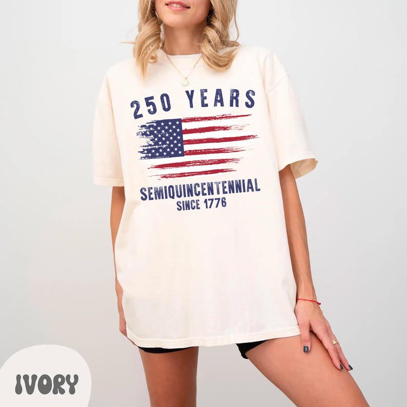 May include: Ivory-colored t-shirt with a distressed American flag graphic. The shirt features the text "250 YEARS" above the flag and "SEMIQUINCENTENNIAL SINCE 1776" below. The shirt is a relaxed fit.