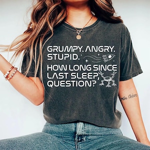 May include: Dark gray t-shirt with white text: "GRUMPY. ANGRY. STUPID. HOW LONG SINCE LAST SLEEP, QUESTION?" Features a rocket ship and a whimsical creature graphic. The shirt is a casual wear item.