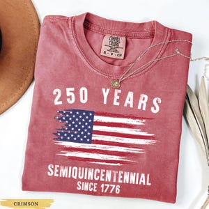 May include: Crimson-colored t-shirt with the text "250 YEARS" above a distressed American flag graphic. Below the flag, the text reads "SEMIQUINCENTENNIAL SINCE 1776". A silver necklace with a pendant is draped across the shirt.