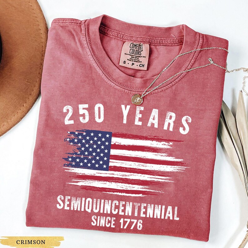 May include: Crimson-colored t-shirt with the text "250 YEARS" above a distressed American flag graphic. Below the flag, the text reads "SEMIQUINCENTENNIAL SINCE 1776". A silver necklace with a pendant is draped across the shirt.