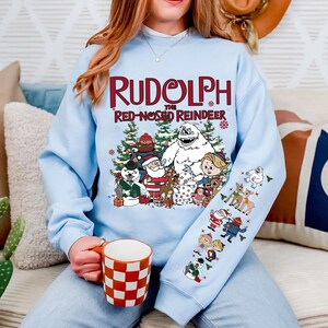 May include: Light blue sweatshirt featuring the text "Rudolph the Red-Nosed Reindeer" with a graphic of characters from the classic Christmas special. The sleeve has additional character graphics. A red and white checkered mug is held in the hand.