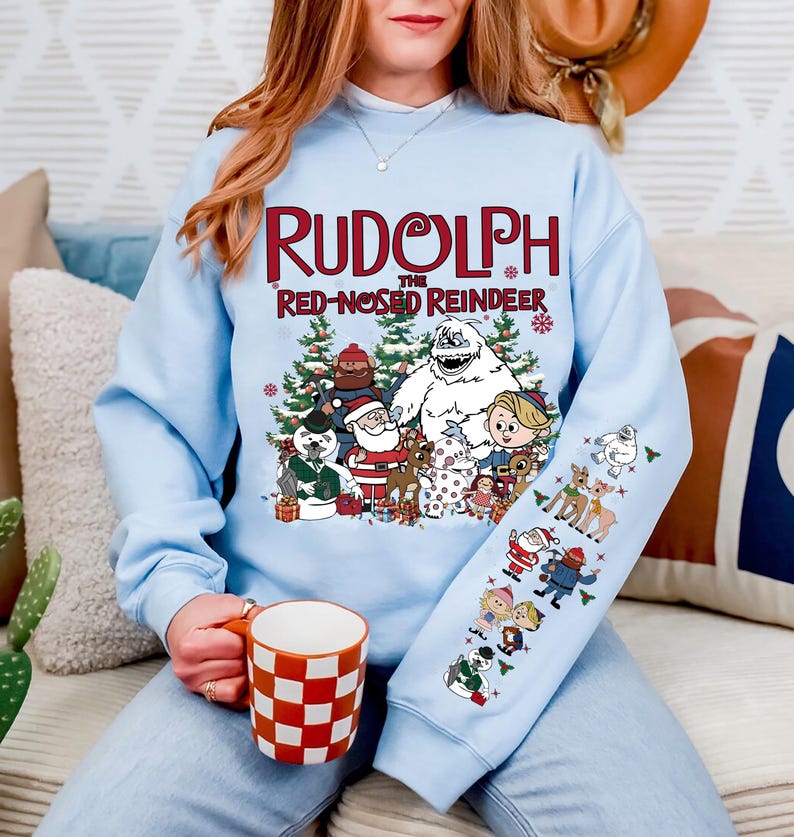 May include: Light blue sweatshirt featuring the text "Rudolph the Red-Nosed Reindeer" with a graphic of characters from the classic Christmas special. The sleeve has additional character graphics. A red and white checkered mug is held in the hand.