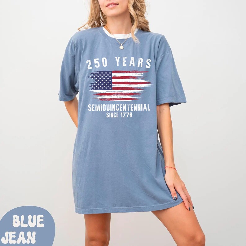 May include: A blue jean colored t-shirt with the text "250 YEARS" above a stylized American flag graphic. Below the flag, the text reads "SEMIQUINCENTENNIAL SINCE 1776". The shirt has a white collar.