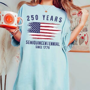 May include: Light blue t-shirt with the text "250 YEARS" above a distressed American flag graphic. Below the flag, the shirt reads "SEMIQUINCENTENNIAL SINCE 1776". A white and orange mug with a sun design is held in the left hand.