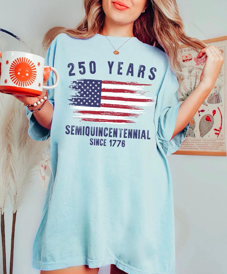 May include: Light blue t-shirt with the text "250 YEARS" above a distressed American flag graphic. Below the flag, the shirt reads "SEMIQUINCENTENNIAL SINCE 1776". A white and orange mug with a sun design is held in the left hand.