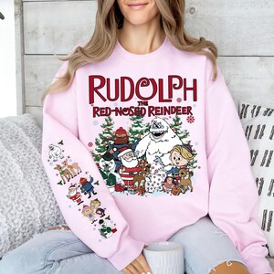 May include: Pink sweatshirt featuring the text "Rudolph the Red-Nosed Reindeer" with cartoon characters. The design includes Santa Claus, a snowman, and reindeer. The left sleeve has additional cartoon illustrations.
