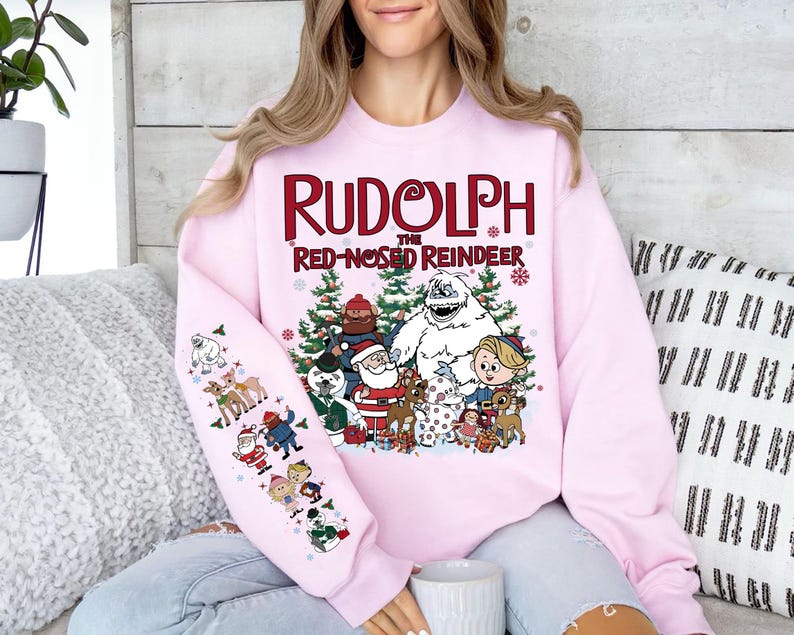 May include: Pink sweatshirt featuring the text "Rudolph the Red-Nosed Reindeer" with cartoon characters. The design includes Santa Claus, a snowman, and reindeer. The left sleeve has additional cartoon illustrations.