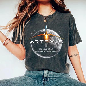 Pode incluir: Camiseta cinza escuro com o logotipo Artemis II, um foguete lançando de um planeta e o texto "We have liftoff". A data e hora do lançamento também são impressas na camiseta de gola redonda.