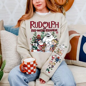 May include: Beige sweatshirt featuring the text "Rudolph the Red-Nosed Reindeer" above a graphic of characters from the classic Christmas special. The sleeve has additional character illustrations. A red and white checkered mug is held in the person's hand.