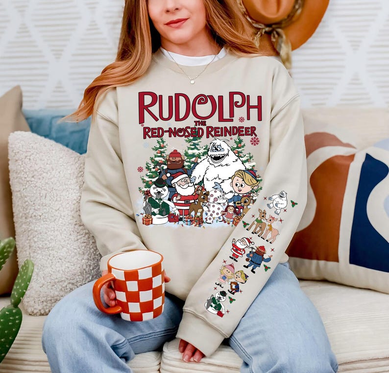 May include: Beige sweatshirt featuring the text "Rudolph the Red-Nosed Reindeer" above a graphic of characters from the classic Christmas special. The sleeve has additional character illustrations. A red and white checkered mug is held in the person's hand.