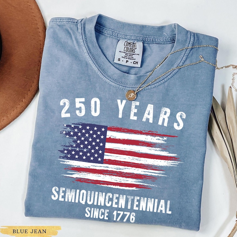 May include: Blue jean colored t-shirt with the text "250 YEARS" above a stylized American flag graphic. Below the flag, the text reads "SEMIQUINCENTENNIAL SINCE 1776". A gold necklace with a pendant is draped on the shirt.
