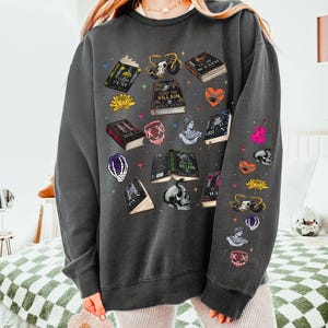 May include: Dark gray sweatshirt with a variety of book and skull graphics. The sweatshirt features illustrations of open books, skulls, hearts, and other gothic-themed designs. The text "HUNT THE VILLAIN" is visible on one of the books.