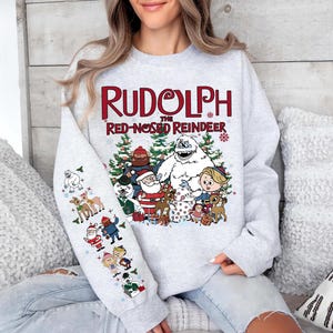 May include: A light gray sweatshirt featuring the text "Rudolph the Red-Nosed Reindeer" in red and white. The design includes cartoon characters from the classic Christmas special, including Rudolph, Santa, and the Abominable Snowman.