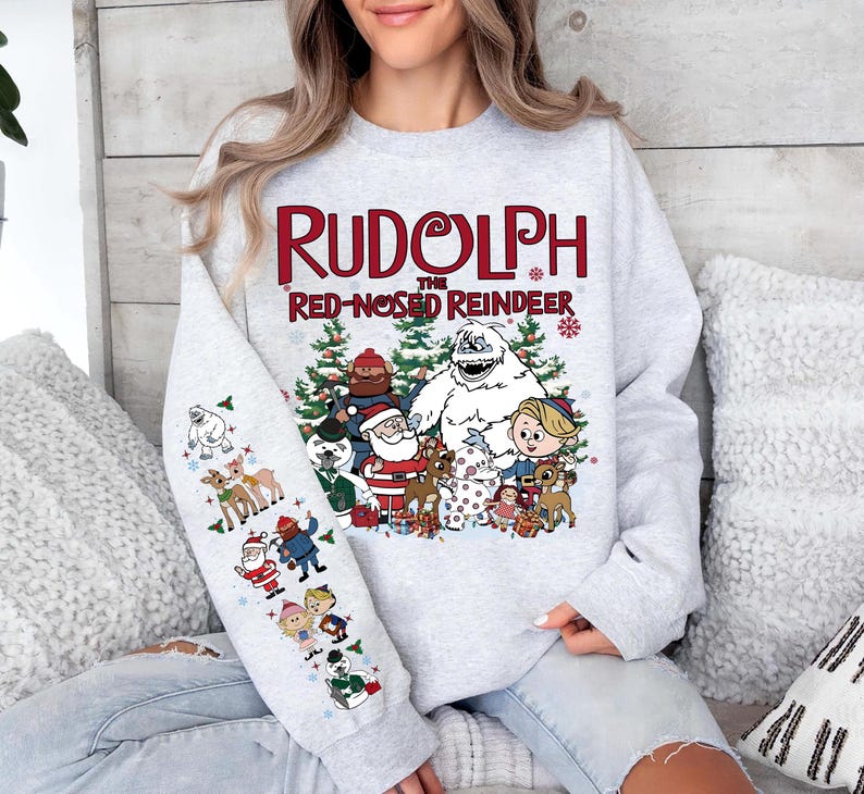 May include: A light gray sweatshirt featuring the text "Rudolph the Red-Nosed Reindeer" in red and white. The design includes cartoon characters from the classic Christmas special, including Rudolph, Santa, and the Abominable Snowman.