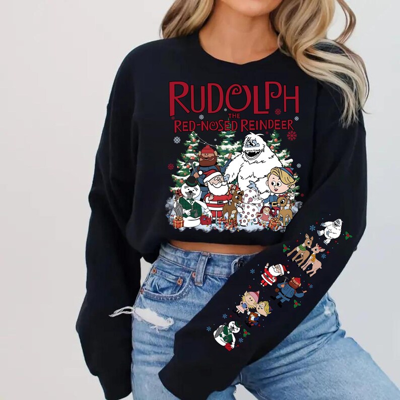 May include: Black sweatshirt featuring a colorful graphic of characters from "Rudolph the Red-Nosed Reindeer." The design includes Santa Claus, a snowman, and other figures. The sleeve also has smaller character illustrations.