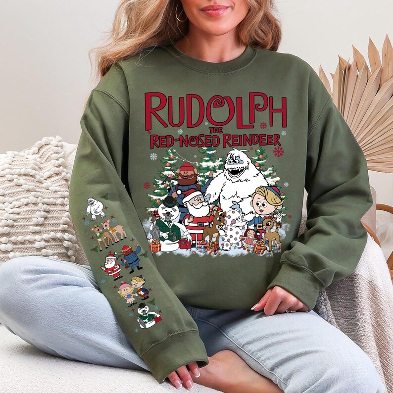 May include: Olive green sweatshirt featuring the text "RUDOLPH the RED-NOSED REINDEER" in red. The design includes cartoon characters from the classic Christmas special. The left sleeve has additional character graphics.