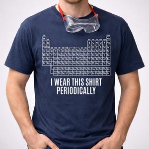 Puede incluir: Camiseta azul marino con la tabla periódica de los elementos y el texto "I WEAR THIS SHIRT PERIODICALLY" en blanco. Gafas de seguridad transparentes con una correa roja alrededor del cuello. Un artículo de ropa con temática científica.