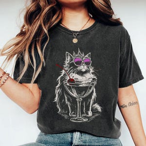 May include: Dark gray t-shirt featuring a cat wearing sunglasses and a crown, sitting next to a martini glass with a cherry. The graphic is in white and black. The person is wearing blue jeans and white shoes.