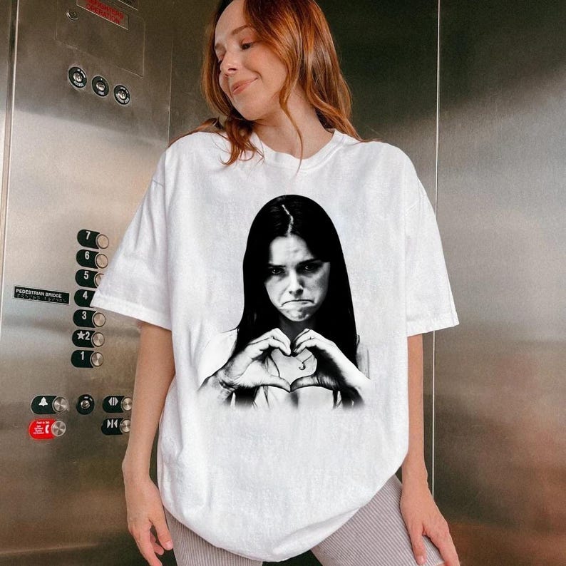 May include: A white short-sleeved t-shirt featuring a black and white graphic print of a person with long dark hair making a heart shape with their hands. The person has a neutral facial expression and is centered on the front of the garment.