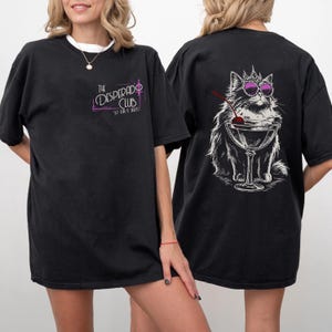 May include: Black t-shirt with a white crew neck collar. The front features the text "The Desperado Club" in a neon-style font. The back displays a cat wearing sunglasses and a crown, sitting in a martini glass with a cherry.