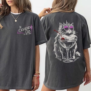 May include: Dark gray t-shirt with a front chest print that reads "The Desperado Club" in a stylized font. The back features a graphic of a cat wearing sunglasses and a crown, with a cocktail glass and cherry.