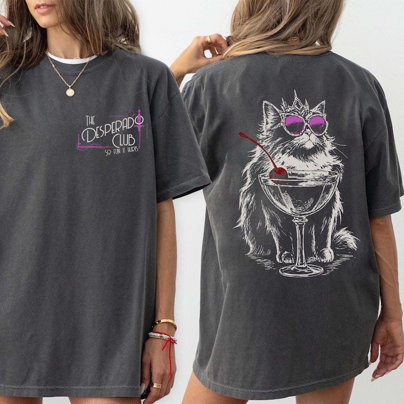 May include: Dark gray t-shirt with a front chest print that reads "The Desperado Club" in a stylized font. The back features a graphic of a cat wearing sunglasses and a crown, with a cocktail glass and cherry.
