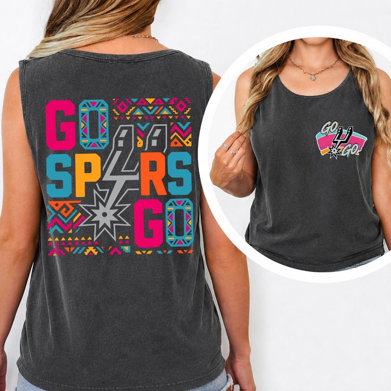 Puede incluir: Una camiseta sin mangas de color gris carb&oacute;n con un gr&aacute;fico grande en la espalda con el texto GO SPURS GO en rosa brillante, naranja y azul, acentuado por patrones geom&eacute;tricos y un logo de espuela plateado. Un logo m&aacute;s peque&ntilde;o est&aacute; en el pecho.