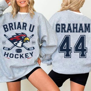 May include: A light gray crewneck sweatshirt featuring a Briar U Hockey graphic with a cardinal bird mascot, hockey sticks, and a puck on the front. The back displays the name Graham and the number 44 in large dark blue block lettering.