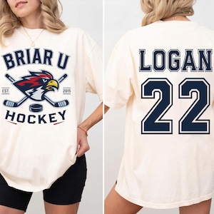 May include: An off-white short-sleeve t-shirt featuring a Briar U Hockey graphic on the front with a cardinal bird mascot, hockey sticks, and a puck. The back of the shirt displays the name Logan and the number 22 in large dark blue block lettering.