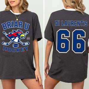 May include: A dark charcoal gray short-sleeve t-shirt. The front features a Briar U Hockey logo with a hawk, hockey sticks, and a puck. The back displays the name Di Laurentis and the number 66 in large blue font with white outlines.