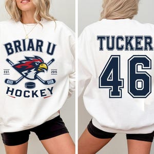 May include: A white crewneck sweatshirt featuring a front graphic of a red and blue bird mascot with two crossed hockey sticks and a puck, with the text BRIAR U HOCKEY EST 2015. The back displays the name TUCKER and the number 46 in navy blue.