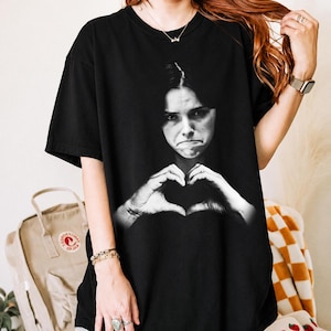 May include: A black short-sleeve t-shirt featuring a large, centered black and white photographic print of a person with a serious expression making a heart shape with their hands. The shirt has a crew neckline and a relaxed, loose-fitting silhouette.
