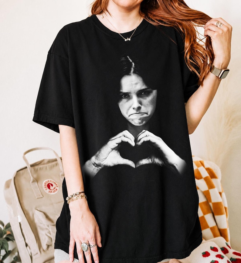 May include: A black short-sleeve t-shirt featuring a large, centered black and white photographic print of a person with a serious expression making a heart shape with their hands. The shirt has a crew neckline and a relaxed, loose-fitting silhouette.