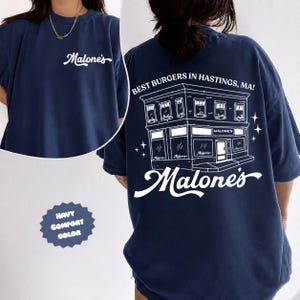 May include: Navy blue comfort color t-shirt with the word "Malone's" in white script on the front. The back features a white illustration of a building with the text "BEST BURGERS IN HASTINGS, MA!" and the name "Malone's".