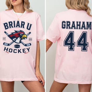 May include: A light pink short-sleeve t-shirt featuring a graphic on the front with the text BRIAR U HOCKEY, EST. 2015, and a hawk mascot with crossed hockey sticks. The back of the shirt displays the name GRAHAM and the number 44 in navy blue.