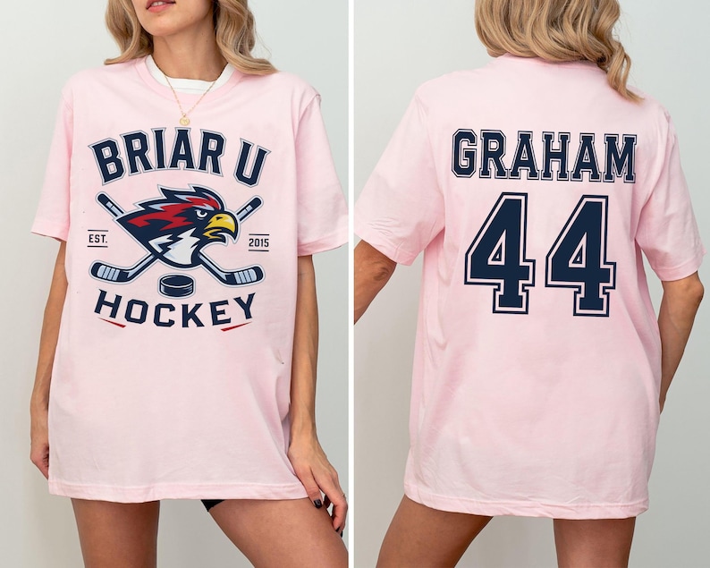 May include: A light pink short-sleeve t-shirt featuring a graphic on the front with the text BRIAR U HOCKEY, EST. 2015, and a hawk mascot with crossed hockey sticks. The back of the shirt displays the name GRAHAM and the number 44 in navy blue.