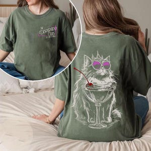 May include: Olive green t-shirt with a graphic of a cat wearing sunglasses and a crown, holding a cocktail with a cherry. The front of the shirt has the text "The Desperado Club" in pink and purple.