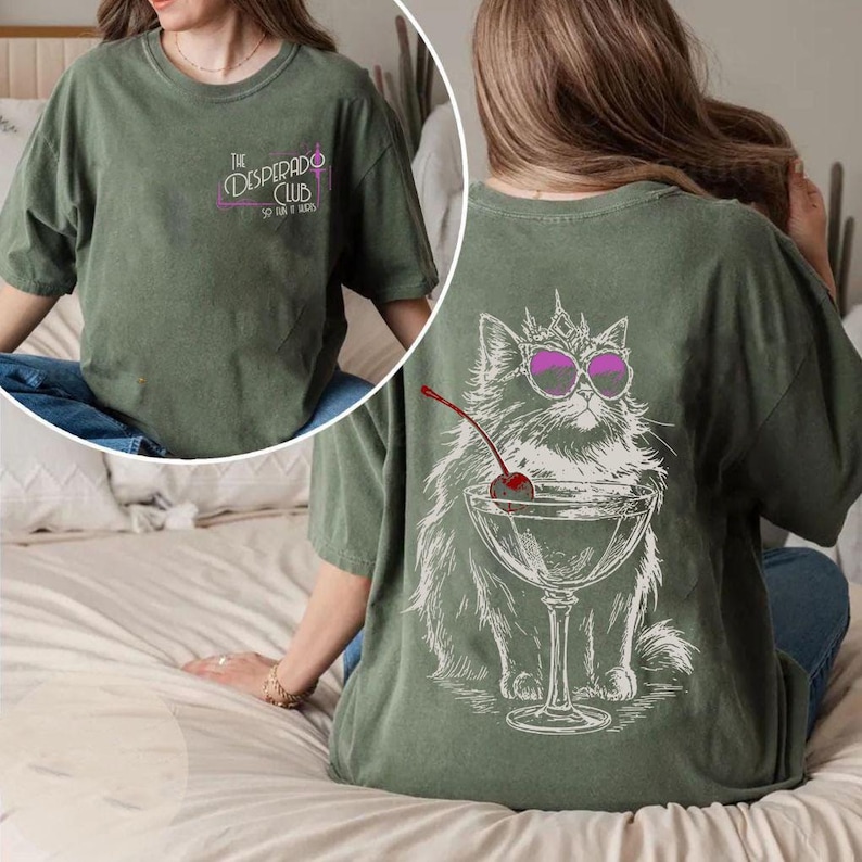 May include: Olive green t-shirt with a graphic of a cat wearing sunglasses and a crown, holding a cocktail with a cherry. The front of the shirt has the text "The Desperado Club" in pink and purple.