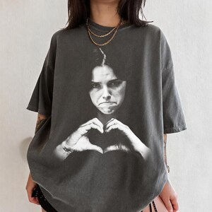 May include: A charcoal gray short-sleeve t-shirt featuring a large, black and white photographic print of a person with a serious expression making a heart shape with their hands. The shirt has a crew neckline and a relaxed, oversized fit.