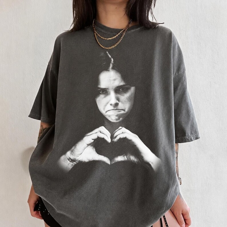 May include: A charcoal gray short-sleeve t-shirt featuring a large, black and white photographic print of a person with a serious expression making a heart shape with their hands. The shirt has a crew neckline and a relaxed, oversized fit.