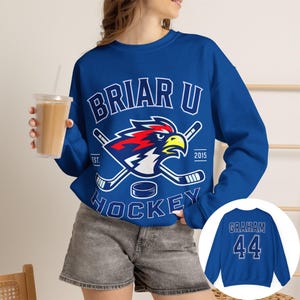 May include: A royal blue crewneck sweatshirt featuring a white graphic of a hawk head, two crossed hockey sticks, and a puck. The text reads BRIAR U HOCKEY EST. 2015. The back of the sweatshirt displays the name GRAHAM and the number 44 in white.