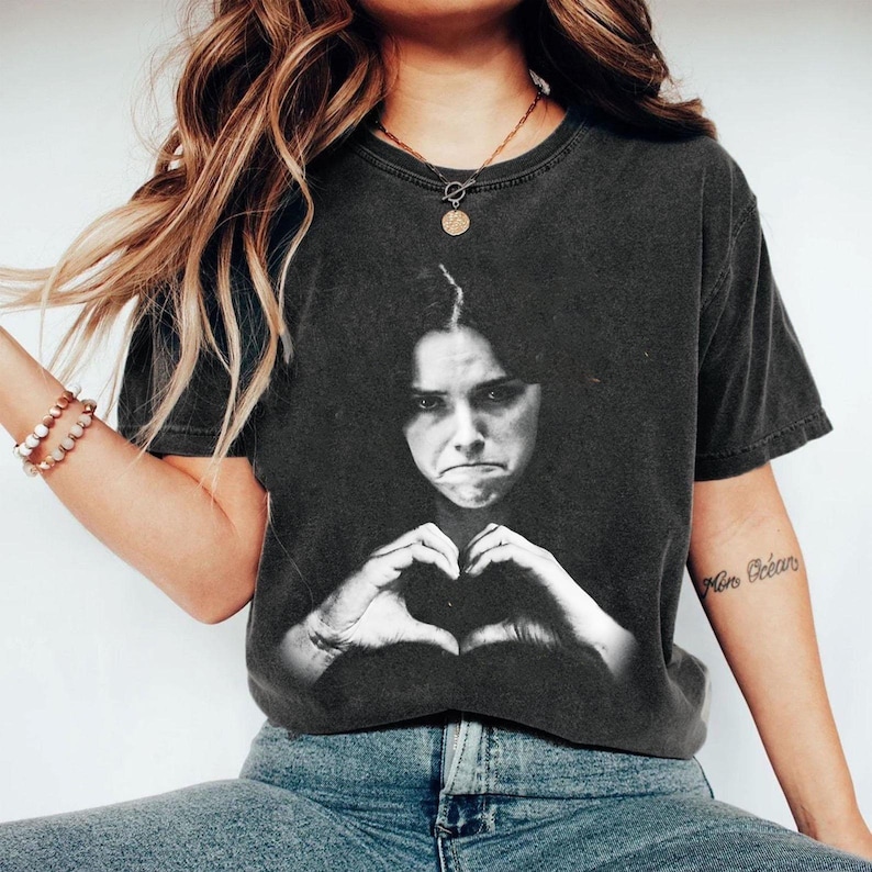 May include: A charcoal gray short-sleeve t-shirt featuring a black and white photographic print of a person with a somber expression making a heart shape with their hands. The shirt has a crew neckline and a faded, vintage-inspired wash finish.