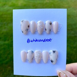 May include: Two sets of press-on nails on a white card. The nails are a pale pink with spiderweb and spider designs. The text "@whimandroot" is written in blue. A hand with a gold and pearl nail design is in the foreground.