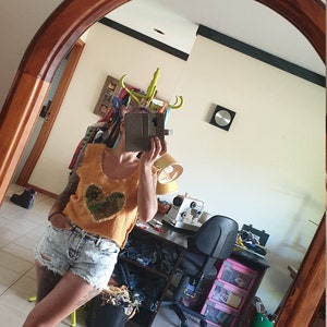 May include: A woman wearing a yellow tank top with a heart-shaped patch and light blue denim shorts. She is standing in front of a mirror and holding a phone in her hand.