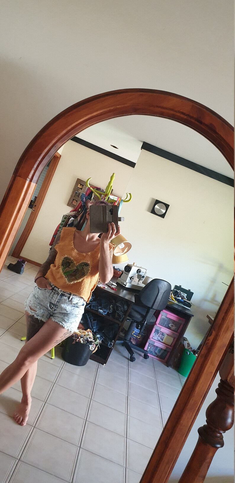 May include: A woman wearing a yellow tank top with a heart-shaped patch and light blue denim shorts. She is standing in front of a mirror and holding a phone in her hand.