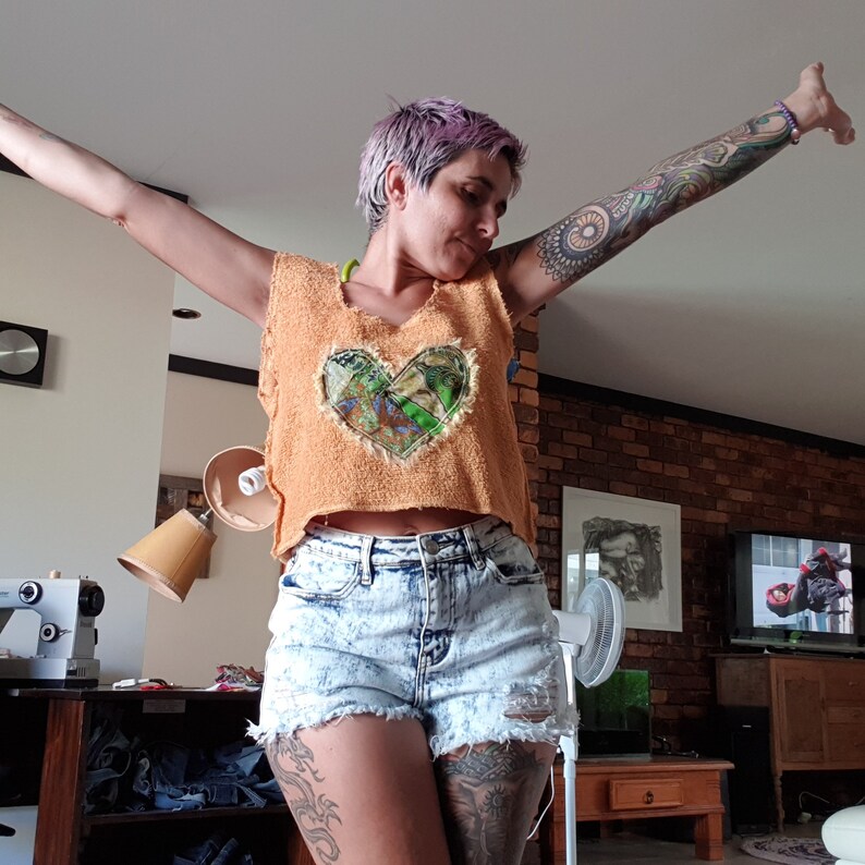 May include: A woman wearing a cropped, orange, fuzzy vest with a heart-shaped patch made of colorful fabric. She is also wearing denim shorts with distressing and a tattoo on her leg.