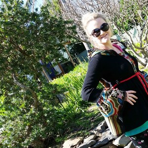 Könnte beinhalten: Eine Frau trägt einen schwarzen Kapuzenpullover mit bunten gemusterten Ärmeln und einem roten Gürtel. Sie steht vor einer Steinmauer und grünem Laub.