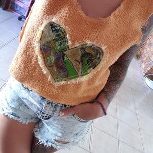 May include: A light orange sleeveless top with a large patchwork heart design. The heart is made of green, brown, and white fabric. The top is made of a soft, fuzzy material.