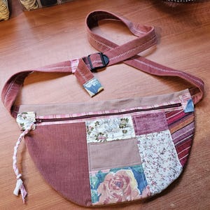 May include: A patchwork fanny pack in shades of mauve, pink, and floral patterns. The bag features a corduroy front panel, a zippered top, and an adjustable strap with a black buckle. A decorative cord hangs from the side.