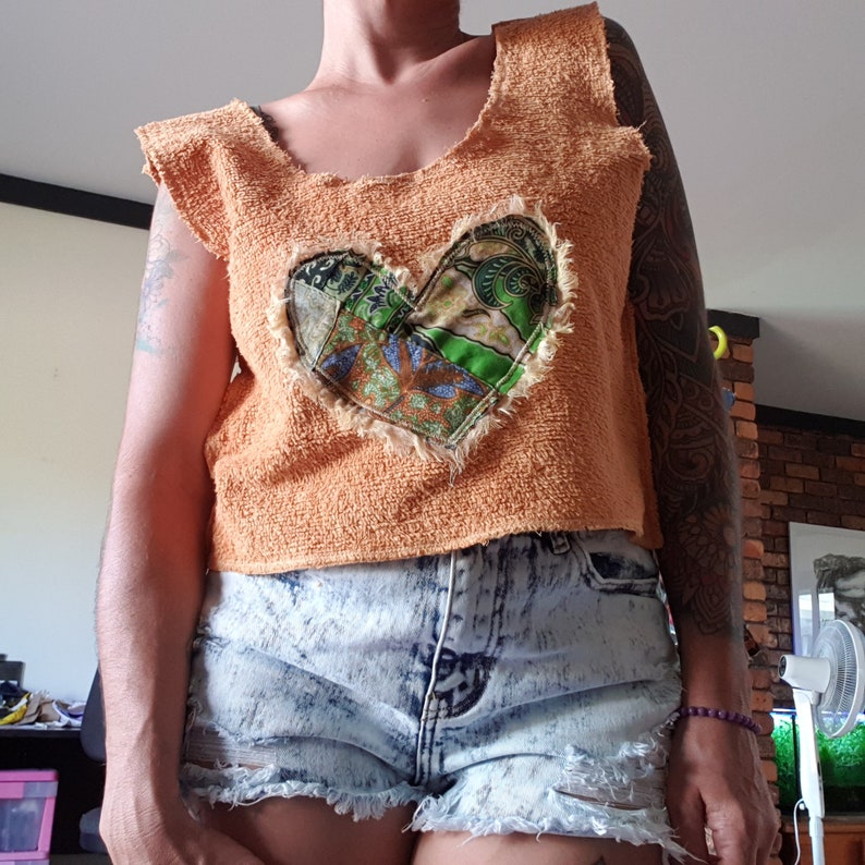 May include: A woman wearing a short-sleeved, orange crocheted top with a patchwork heart design. The top is cropped and the woman is wearing denim shorts.